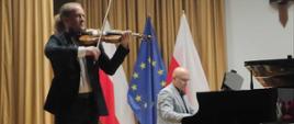 Zdjęcie kolorowe. Dwóch muzyków na znajduje się na scenie. Jeden stoi i gra na skrzypcach. Drugi siedzi przy fortepianie i gra