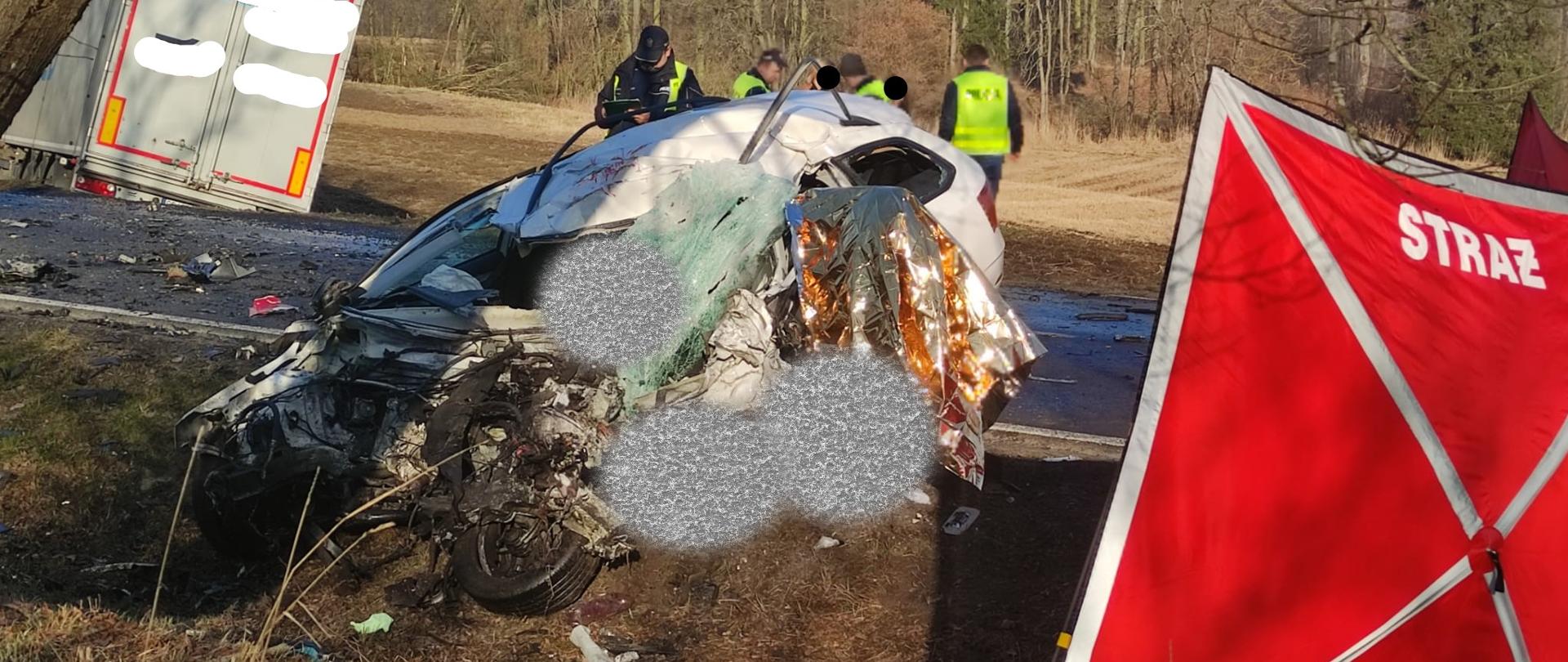 Zdjęcie przedstawia samochód osobowy rozbity w wyniku czołowego zderzenia z samochodem ciężarowym