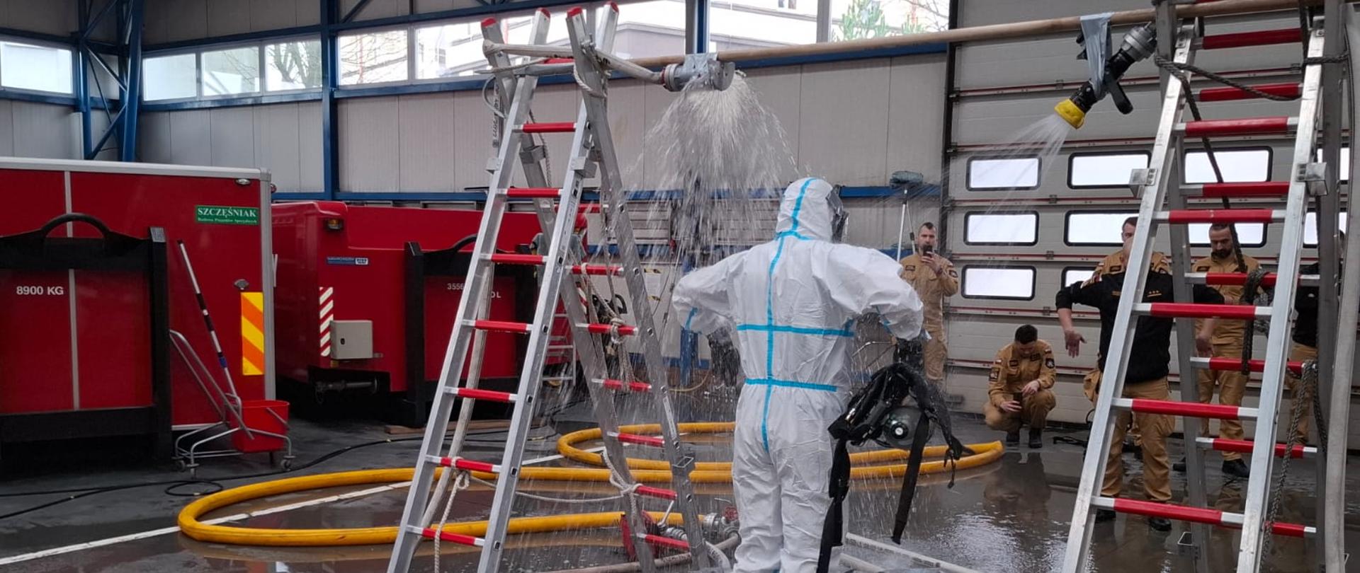 Na zdjęciu widać wnętrze garażu w jednostce Państwowej Straży Pożarnej, w której odbywa się procedura dekontaminacji. Na pierwszym planie stoi strażak ubrany w biały kombinezon ochronny z niebieskimi taśmami uszczelniającymi, czerwonymi gumowymi butami i rękawicami. Osoba ta znajduje się pod prysznicem dekontaminacyjnym – woda leje się na nią z góry, a jej strumień jest rozpraszany przez specjalną dyszę zamontowaną na poziomej rurze. Zdjęcie z warsztatów przedstawia procedurę dekontaminacji, związanej z usuwaniem substancji niebezpiecznych.
Konstrukcja prysznica została zbudowana z dwóch aluminiowych drabin, między którymi umieszczono poziomą rurę z otworami na wodę. Cała scena odbywa się nad specjalną folią zabezpieczającą, tworzącą basen, w którym zbiera się woda. Na podłodze widać liczne węże strażackie, niektóre są podłączone do instalacji wodnej.
W tle kilku strażaków ubranych w piaskowe mundury obserwuje proces. Część z nich stoi, inni siedzą lub kucają, przyglądając się działaniom. W głębi hali znajdują się duże czerwone kontenery z oznaczeniami, prawdopodobnie zawierające sprzęt strażacki i ratowniczy. Na podłodze leżą żółte i szare węże strażackie, które prawdopodobnie doprowadzają wodę do prysznica.