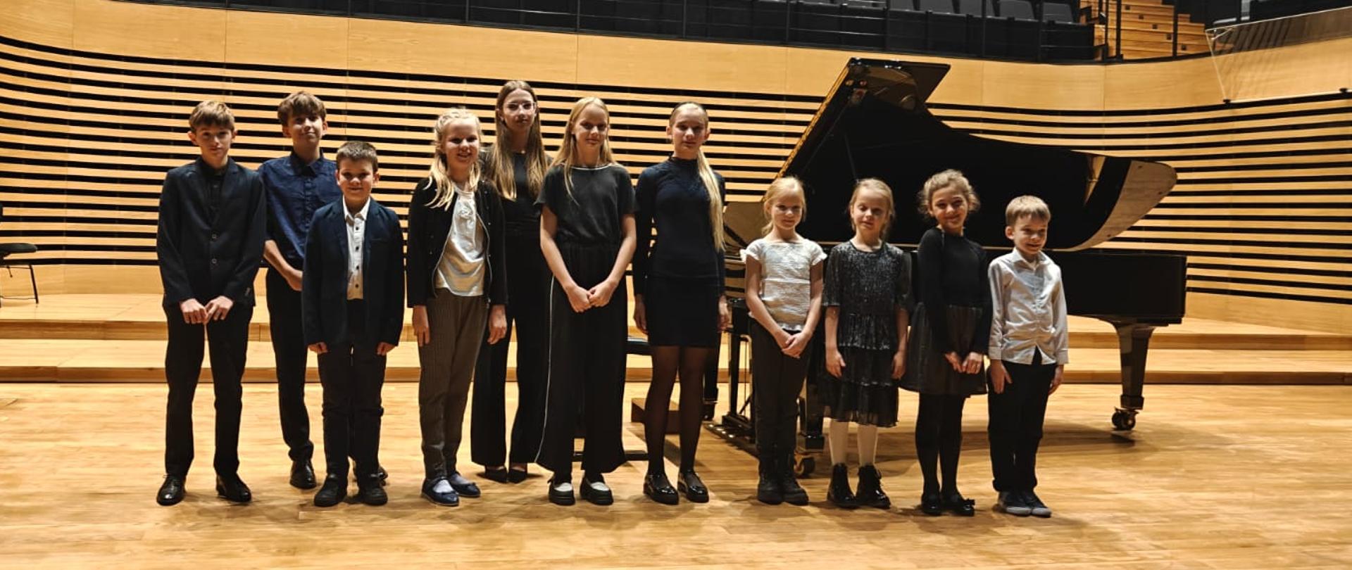 Wszyscy wykonawcy stoją przed fortepianem na estradzie.