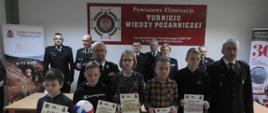 Dzieci stoją ustawione jedno obok drugiego. Trzymają w rękach dyplomy i nagrody. Za nimi stoją strażacy w mundurach wyjściowych. W tle widać baner z logo Ochotniczych Straży Pożarnych i napisem Powiatowe eliminację Turnieju Wiedzy Pożarniczej. Po obu stronach baneru stoją dwa rolapy.