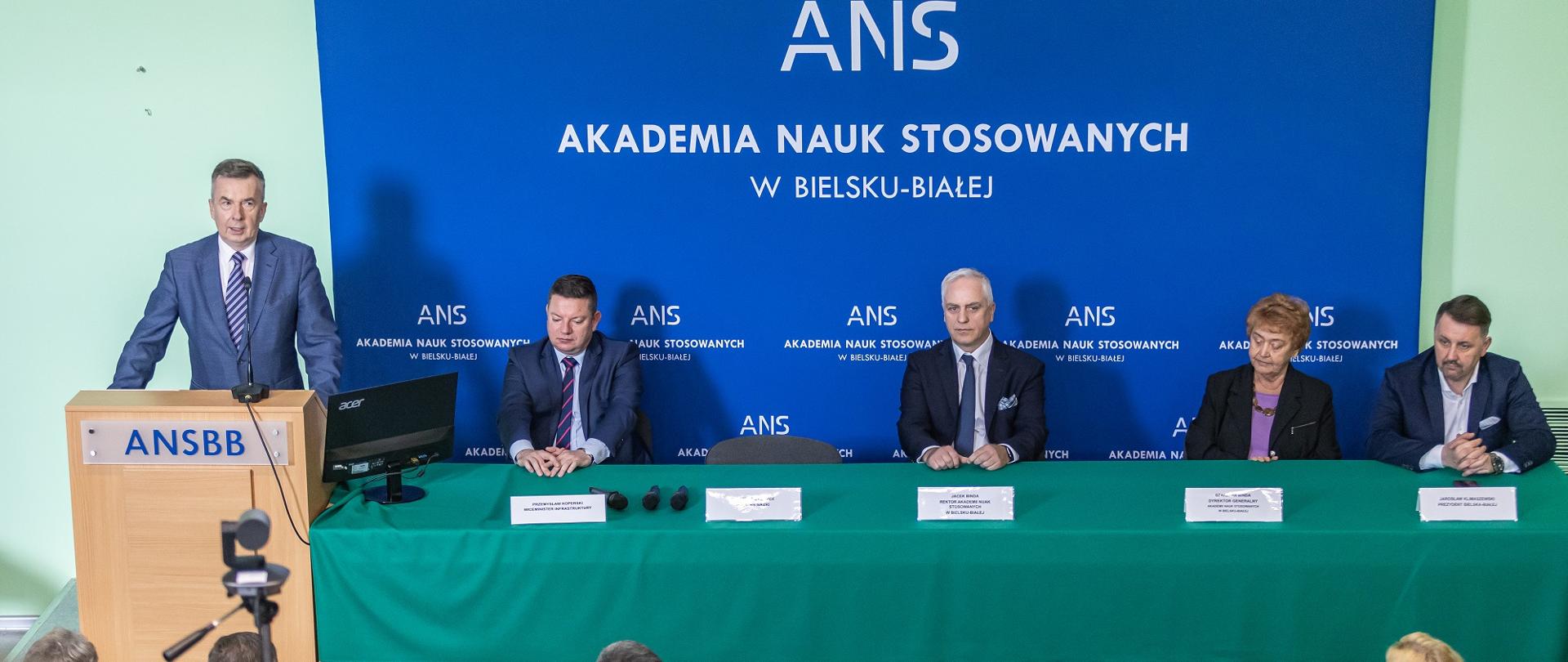 Minister Wieczorek stoi za mównicą i mówi do mikrofonu, za nim na niebieskiej ścianie biały napis ANS - Akademia Nauk Stosowanych w Bielsku-Białej, obok ministra za przykrytym zielonym materiałem stołem siedzą cztery osoby.