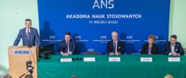 Minister Wieczorek stoi za mównicą i mówi do mikrofonu, za nim na niebieskiej ścianie biały napis ANS - Akademia Nauk Stosowanych w Bielsku-Białej, obok ministra za przykrytym zielonym materiałem stołem siedzą cztery osoby.