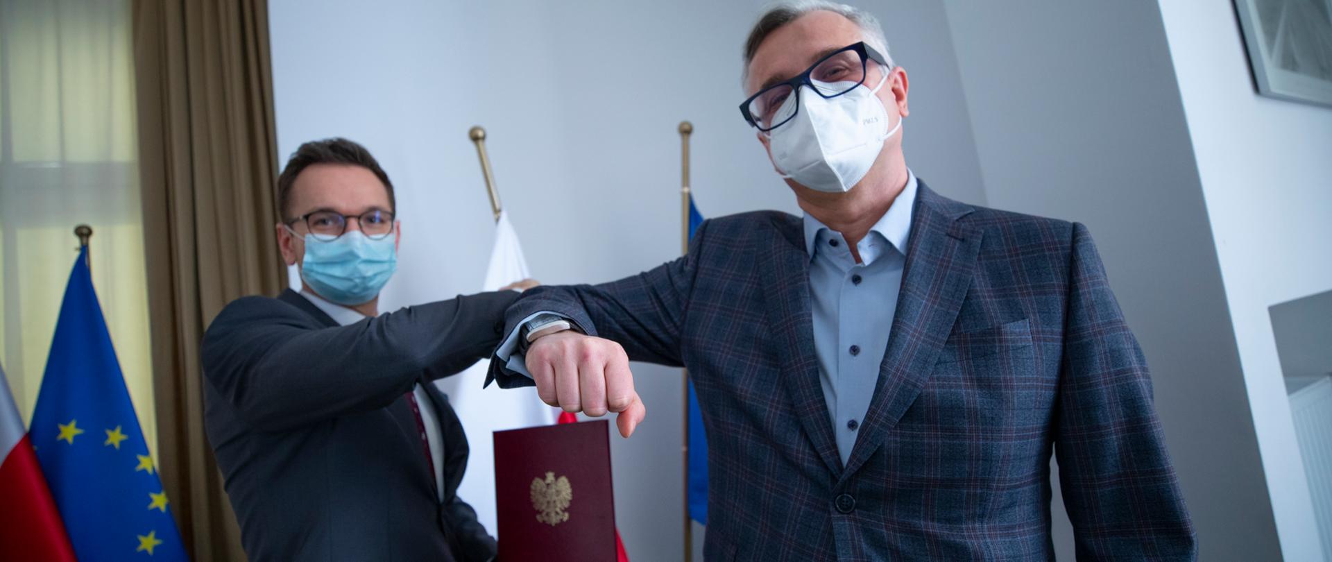 Od lewej wiceminister Waldemar Buda, który trzyma w ręku teczkę z orłem, wita się łokciem z przedstawicielem Centrum Kreatywności i Nowych Technologii.