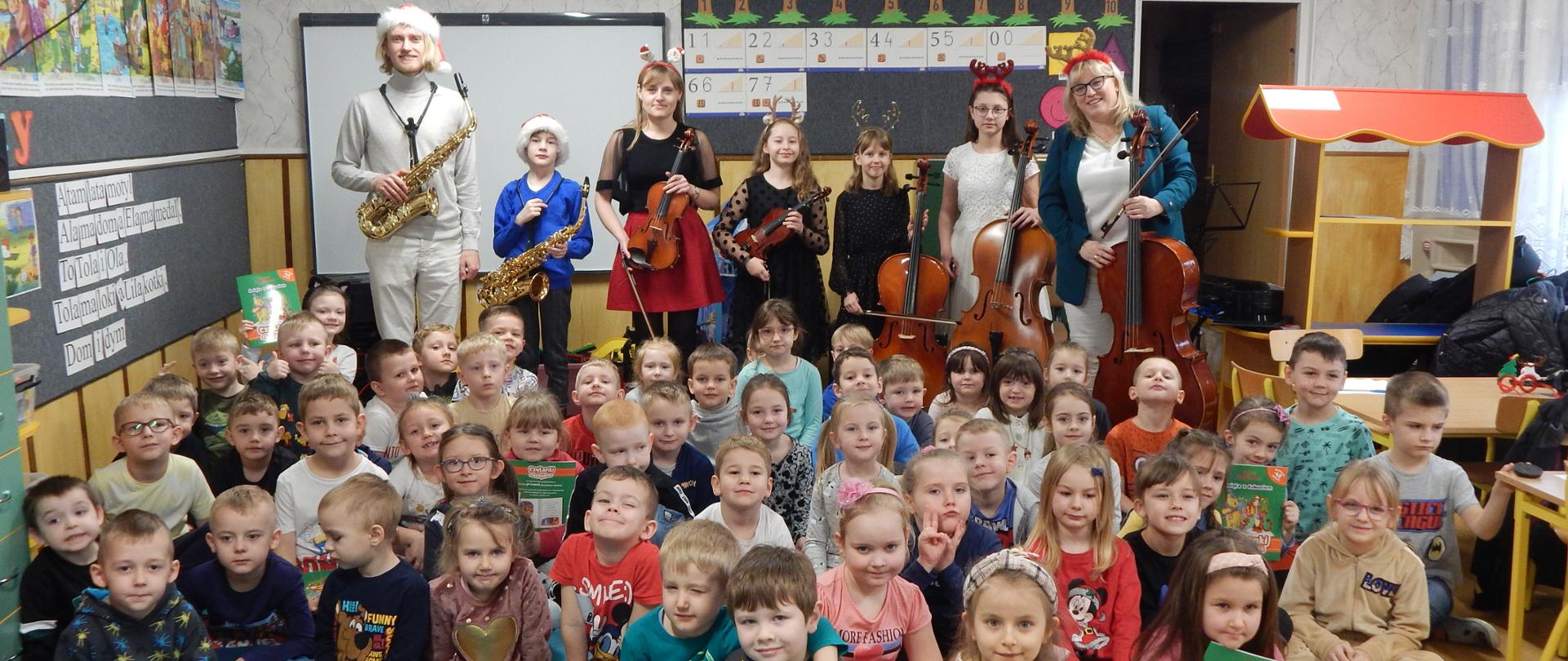 Na zdjęciu sala przedszkolna w Przedszkolu Miejskim nr 4 w Grajewie. Na pierwszym planie dzieci-widzowie siedzący po turecku, za nimi wykonawcy koncertu "Świąteczne smyki i stroiki czyli zabawa z saksofonem, skrzypcami i wiolonczelą". Wykonawcy maja ze sobą instrumenty, wiolonczele, skrzypce, saksofon. Na głowach mają czapki Mikołaja i inne ozdoby świąteczne. W tle po lewej stronie meble i zabawki przedszkolne, na środku monitor interaktywny, oraz tablica, na której zawieszone są wycięte z kolorowego papieru drzewa z namalowanymi jabłkami, pod nimi powieszone obrazki z cyframi, po prawej stronie drewniany stragan-zabawka, stoły i krzesła.