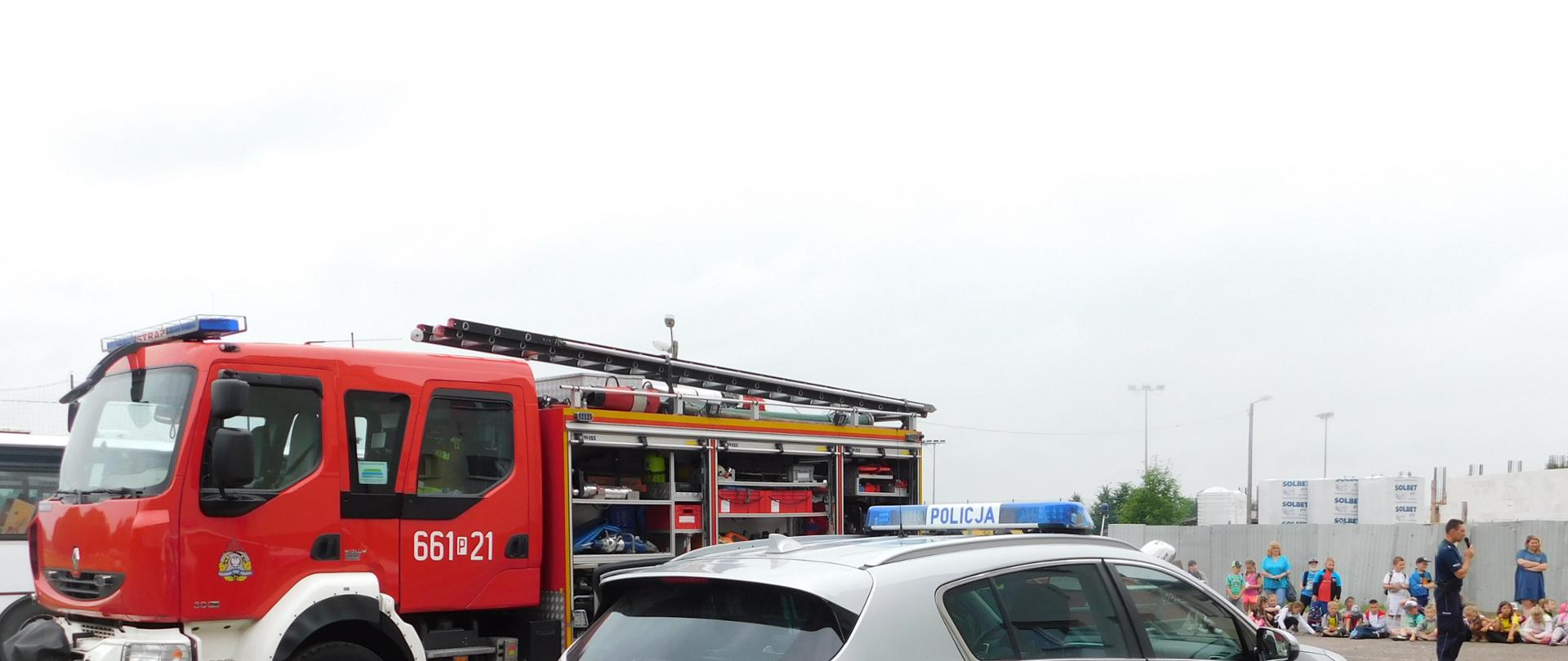 Na zdjęciu widok samochodu strażackiego i policyjnego w tle widać policjanta ,który przekazuje zgromadzonym dzieciom informacje na temat bezpieczeństwa podczas wakacji.