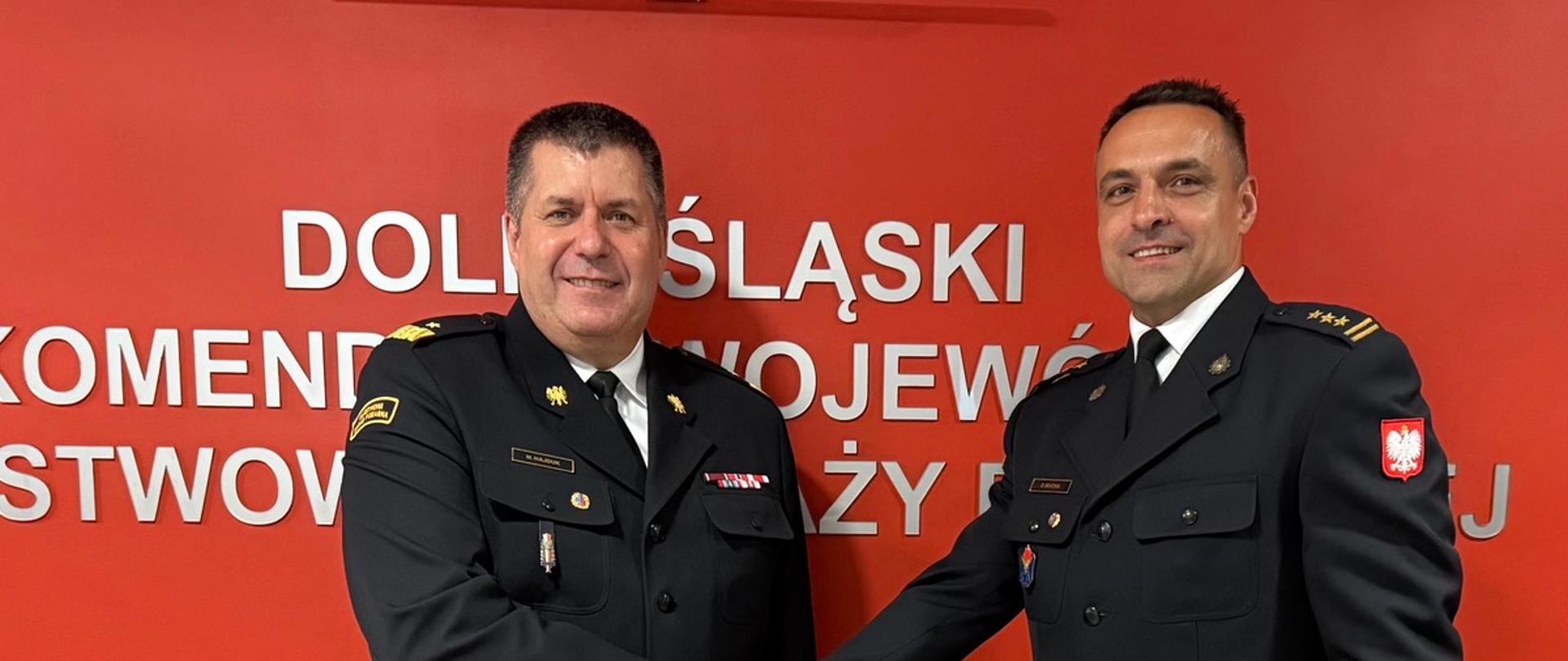 Zdjęcie przedstawia strażaków w ciemnych mundurach galowych Państwowej Straży Pożarnej na tle jaskrawoczerwonej ściany z białym, metalicznym napisem „DOLNOŚLĄSKI KOMENDANT WOJEWÓDZKI PAŃSTWOWEJ STRAŻY POŻARNEJ”