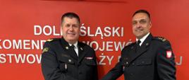 Zdjęcie przedstawia strażaków w ciemnych mundurach galowych Państwowej Straży Pożarnej na tle jaskrawoczerwonej ściany z białym, metalicznym napisem „DOLNOŚLĄSKI KOMENDANT WOJEWÓDZKI PAŃSTWOWEJ STRAŻY POŻARNEJ”
