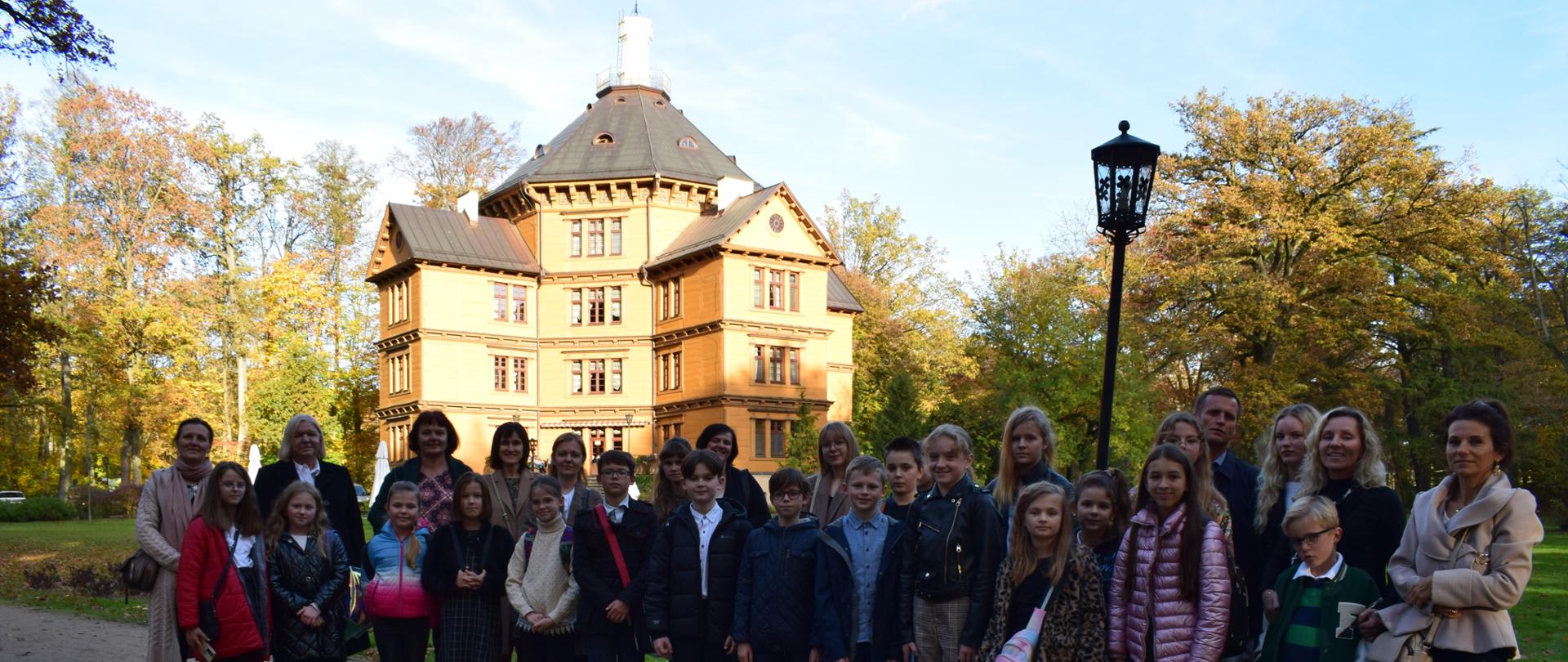 Zdjęcie przedstawia grupę uczniów wraz z opiekunami i rodzicami stojących w dwuszeregu na tle drewnianego Pałacu Myśliwskiego Książąt Radziwiłłów w Antoninie w kształcie ośmioboku z namiotowym dachem i widocznymi dwoma niższymi skrzydłami (dobudówkami), pomalowanego na żółty kolor. Pałac zajmuje centralną część zdjęcia. Po lewej i prawej stronie w tle widać drzewa z kolorowymi liśćmi. Nad Pałacem i drzewami wzdłuż zdjęcia u góry roztacza się jest jasne (lekko błękitne) niebo. Z prawej strony tuż za uczniami stoi wysoka stylowa latarnia.