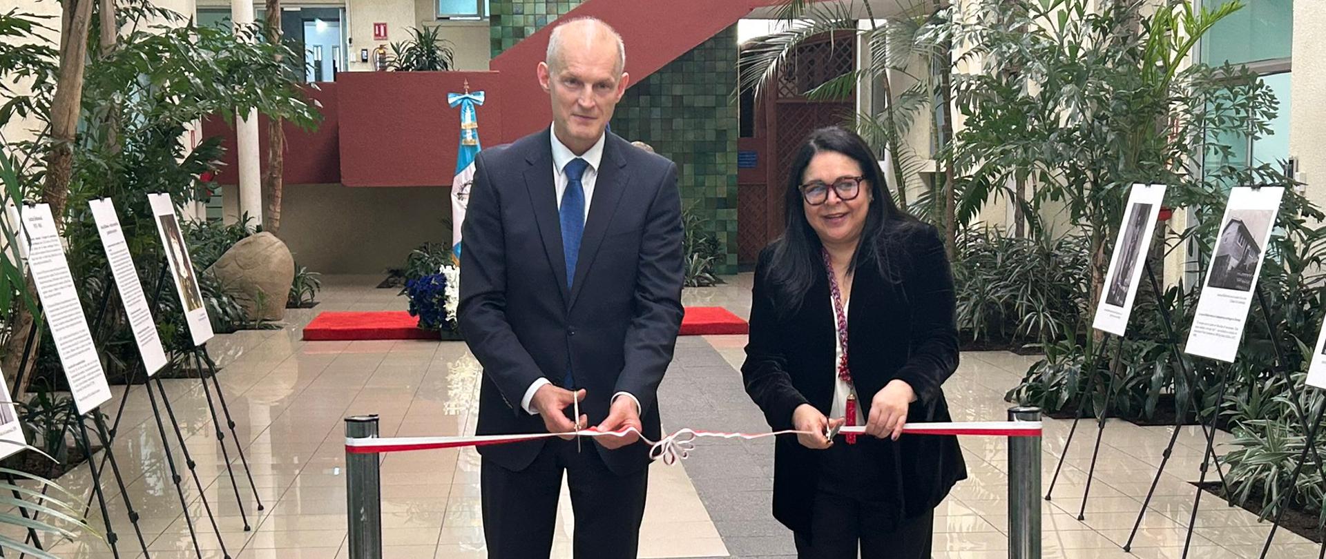 Dwie osoby w formalnych strojach przecinają czerwono-białą wstęgę podczas uroczystego otwarcia wystawy. W tle znajduje się przestronne atrium z roślinami oraz plansze ekspozycyjne ustawione po obu stronach przejścia.