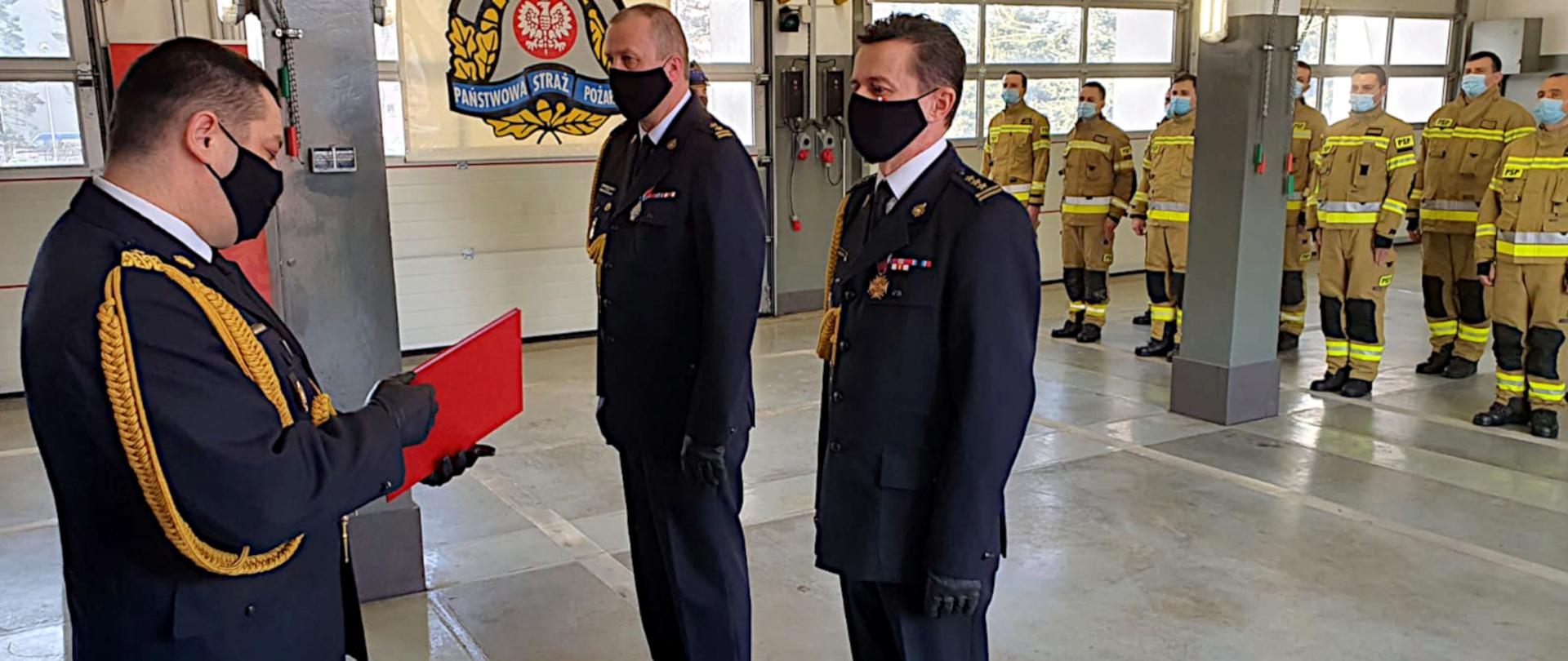 Zdjęcie ilustruje uroczystość powierzenia obowiązków komendanta miejskiego PSP w Jaworznie. W garażu Komendy Miejskiej w Jaworznie z lewej strony stoi w mundurze galowym i maseczce ochronnej Śląski Komendant Wojewódzki PSP nadbryg. Jacek Kleszczewski, który w ręce trzyma akt odwołania. Po prawej stronie ubrani w mundury galowe i maseczki ochronne stoją: odchodzący na zaopatrzenie emerytalne st. bryg. Dariusz Kleszcz i nowo powołany Komendant Miejski PSP w Jaworznie, bryg. Paweł Żaba. Na dalszym planie stoją w dwuszeregu strażacy w ubraniach specjalnych i maseczkach ochronnych.