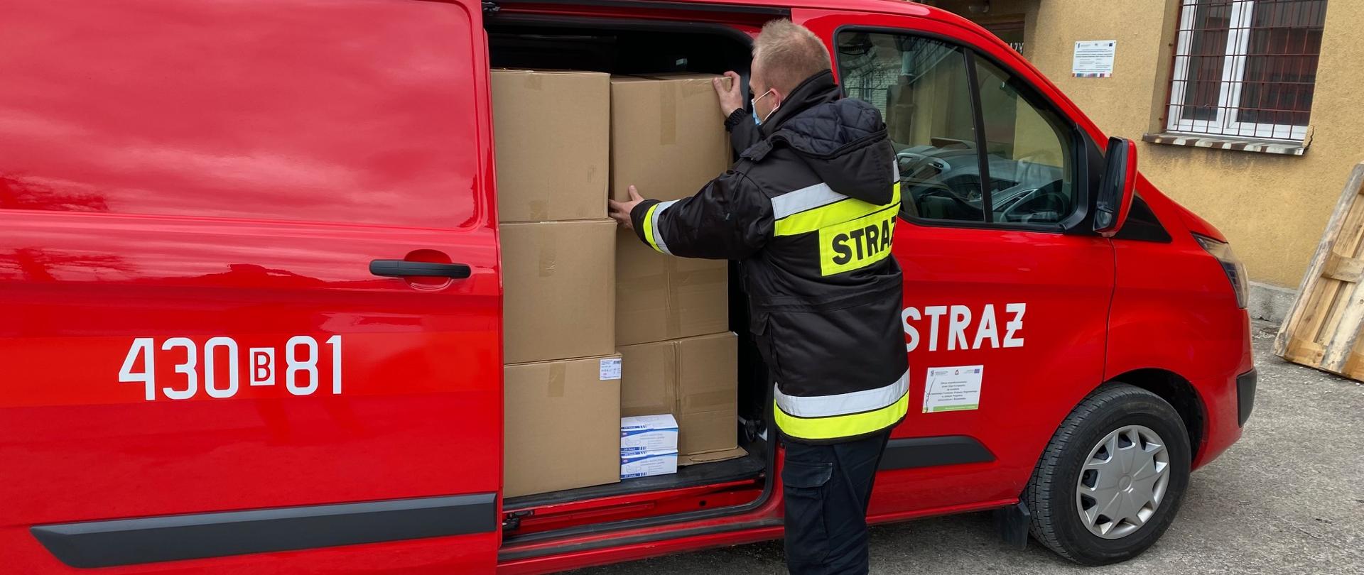 Z czerwonego strażackiego busa strażak wyładowuje kartonowe pudła.