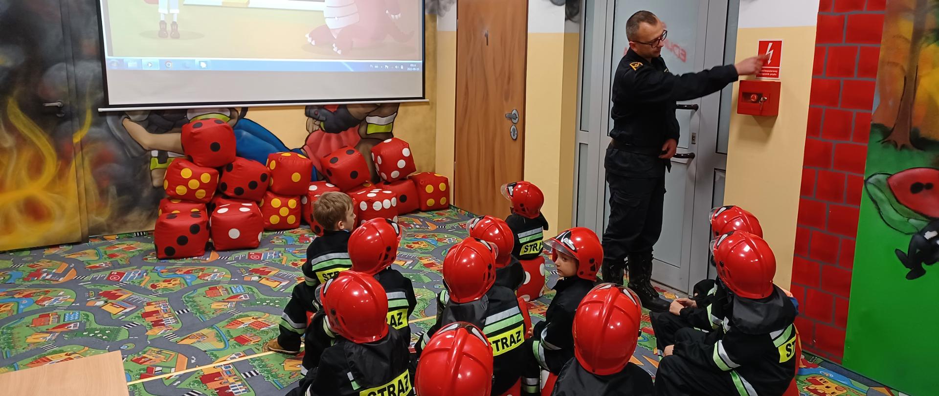 Dzieci w ubrane w przebranie strażackie siedzą na czerwonych pufach w sali edukacyjnej i słuchają strażaka, który prowadzi zajęcia. Na ścianie widoczny jest puszczany filmik z projektora. 
