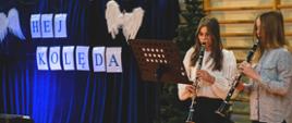 Zdjęcie przedstawia duet klarnetowy złożony z dziewczyn. Są one w trakcie wykonywania utworu muzycznego. Przed nimi stoi wysoki czarny pulpit z nutami. W tle widnieją drabinki gimnastyczne, choinka oraz niebieska kotara ze skrzydłami anioła jako dekoracja i napis "Hej kolęda"