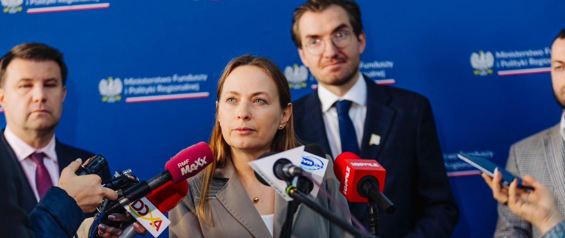 Minister Katarzyna Pełczyńska-Nałęcz stoi z przodu przy mikrofonie.. W tle za nią stoi kilka osób.