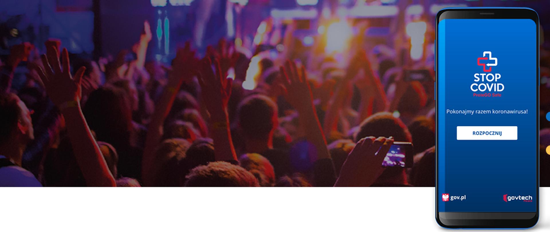 na pierwszym planie smartfon z ekranem startowym aplikacji stop covid w zdjęci tłum ludzi z podniesionymi rękami podczas koncertu