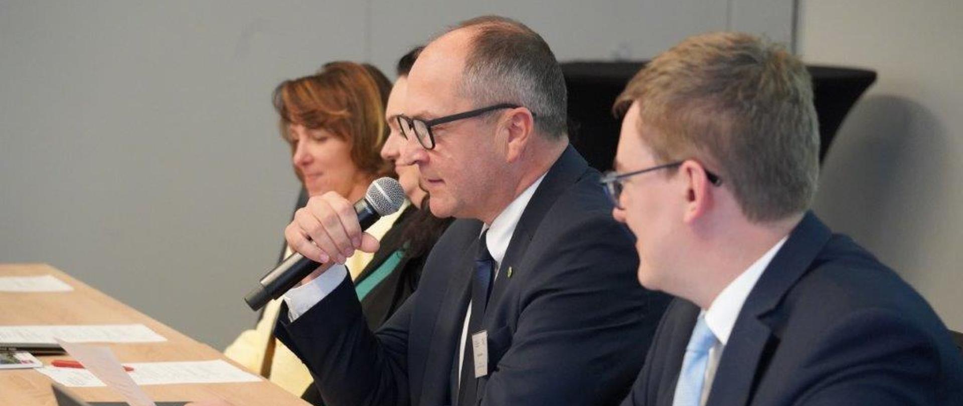 Zbliżenie na dwóch mężczyzn w garniturach siedzących przy stole konferencyjnym. Mężczyzna w okularach pośrodku trzyma mikrofon i mówi, trzymając w drugiej ręce kartkę.