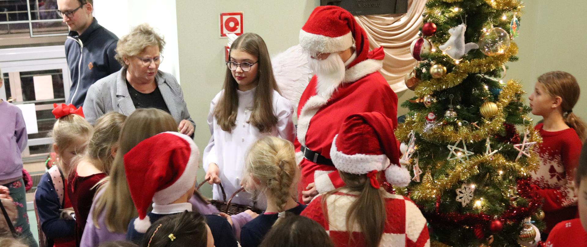 Kolorowe zdjęcie - grupa dzieci ubranych kolorowo z dodatkiem świątecznych akcentów typu czapki Mikołaja. Wszystkie dzieci otaczają Mikołaja i anioła, którzy rozdają im cukierki. Całość odbywa się na tle udekorowanej choinki.