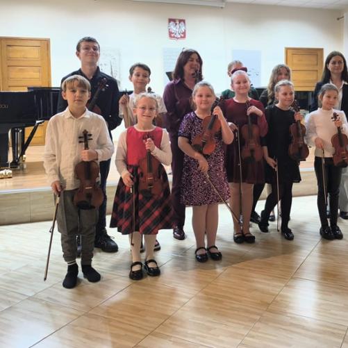 Dzieci stoją ze skrzypcami pod sceną wraz z nauczycielką skrzypiec i akompaniatorką pozując do wspólnego zdjęcia po zakończonej audycji. 