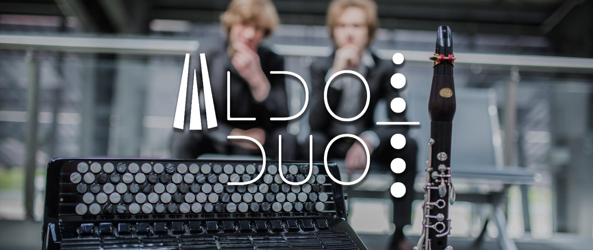 Baner w chłodnej, stonowanej kolorystyce z przewagą szarości i czerni. Na pierwszym planie widoczne są instrumenty: fragment akordeonu z klawiaturą guzikową oraz stojący klarnet. W tle, lekko rozmyci, siedzą dwaj mężczyźni w ciemnych strojach. Centralnie umieszczono białe logo i napis „ALDO DUO” o nowoczesnej, minimalistycznej typografii. Całość ma elegancki, koncertowy charakter.