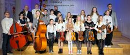 Grupa uczniów oraz pedagodzy w strojach galowych. Uśmiechnięci trzymają w rękach swoje instrumenty. W tle logo szkoły. Ogólna kolorystyka – biało-niebieska.