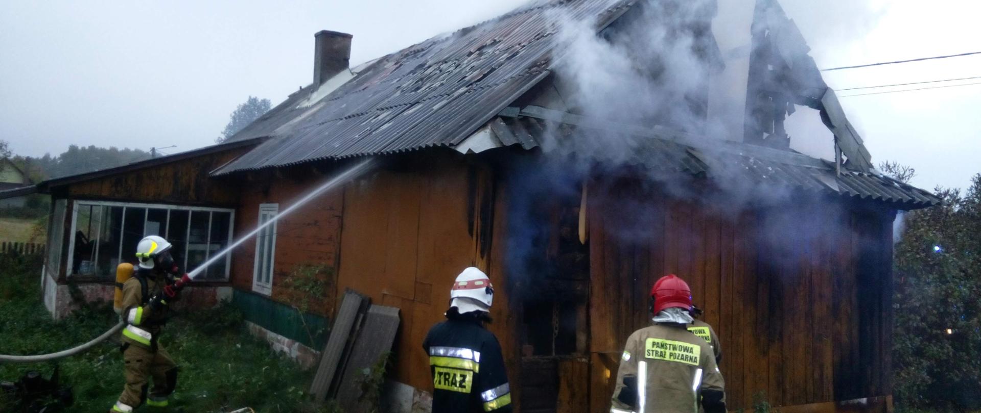 Zdjęcie przedstawia drewniany budynek mieszkalny którego poddasze objęte jest pożarem. Z Okolic dachu wydobywają się kłęby szarego dymu. Do okoła budynku stoją strażacy - jeden z nich ubrany w aparat ochrony dróg oddechowych podaje prąd wody na pożar.
