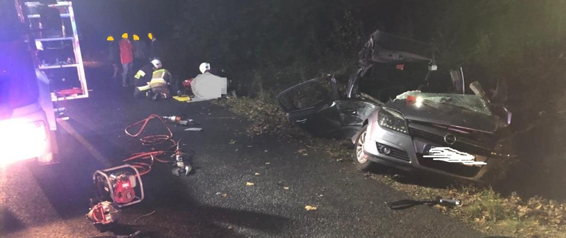 Widok samochodu osobowego po uderzeniu w auto. Ratownicy użyli narzędzi hydraulicznych by wydostać poszkodowanych. W tle widać jak strażacy udzielają pomocy medycznej.