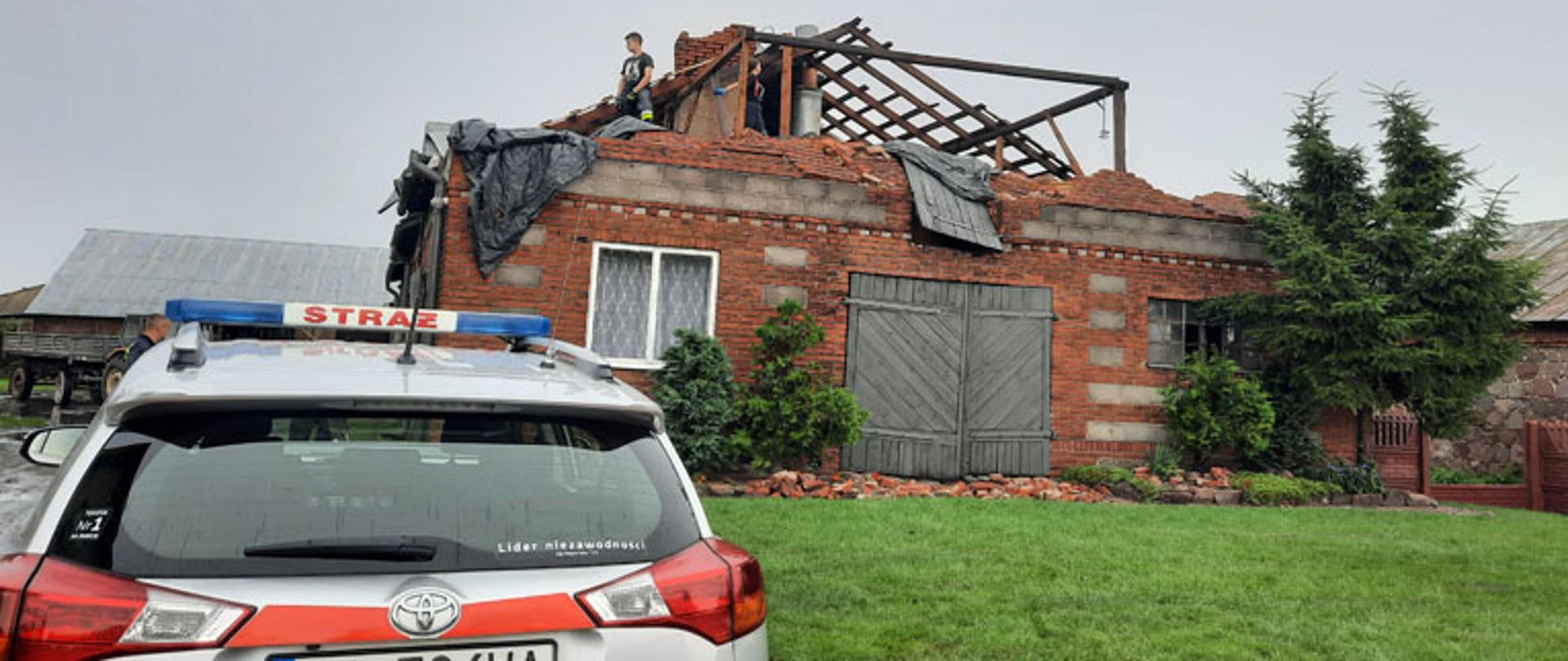 Zdjęcie przedstawia dom jednorodzinny, na zniszczonym dachu dwóch strażaków przykrywa dziurę po zerwanym przez wiatr poszyciu dachowym. Po lewej stronie zdjęcia budynek gospodarczy a przed nim traktor z przyczepą. Na terenie posesji stoi samochód operacyjny straży pożarnej. Niebo ciemnoszare, deszczowe