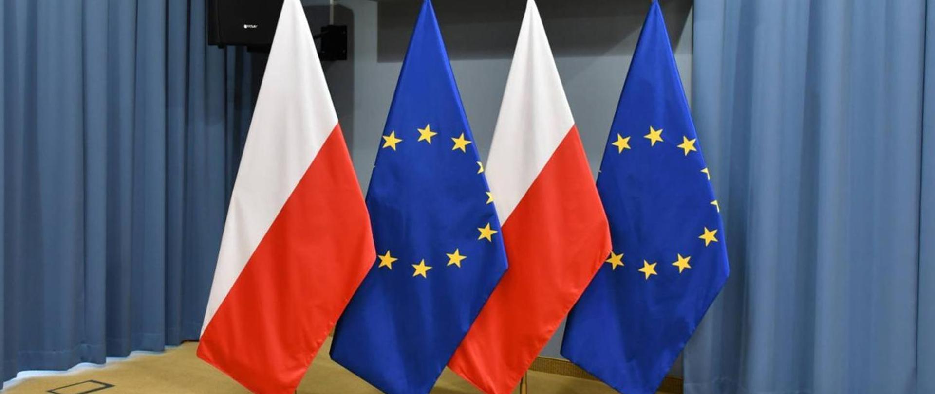 Dwie flagi Polski i dwie flagi Unii Europejskiej na tle niebieskich zasłon.
