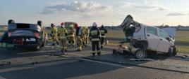 Uszkodzony samochód osobowy leży na dachu. W tle drugi uszkodzony samochód osobowy oraz samochód strażacki i strażacy.