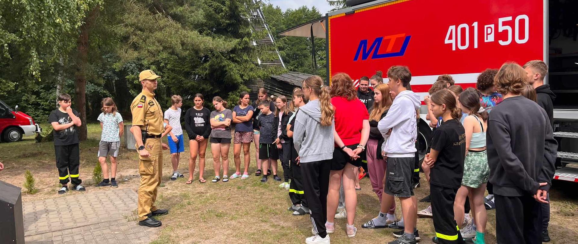 Zdjęcie przedstawia strażaka przeprowadzającego edukację dla dzieci i młodzieży na wakacyjnym obozie strażackim.
W tle świetlica i samochód strażacki.
