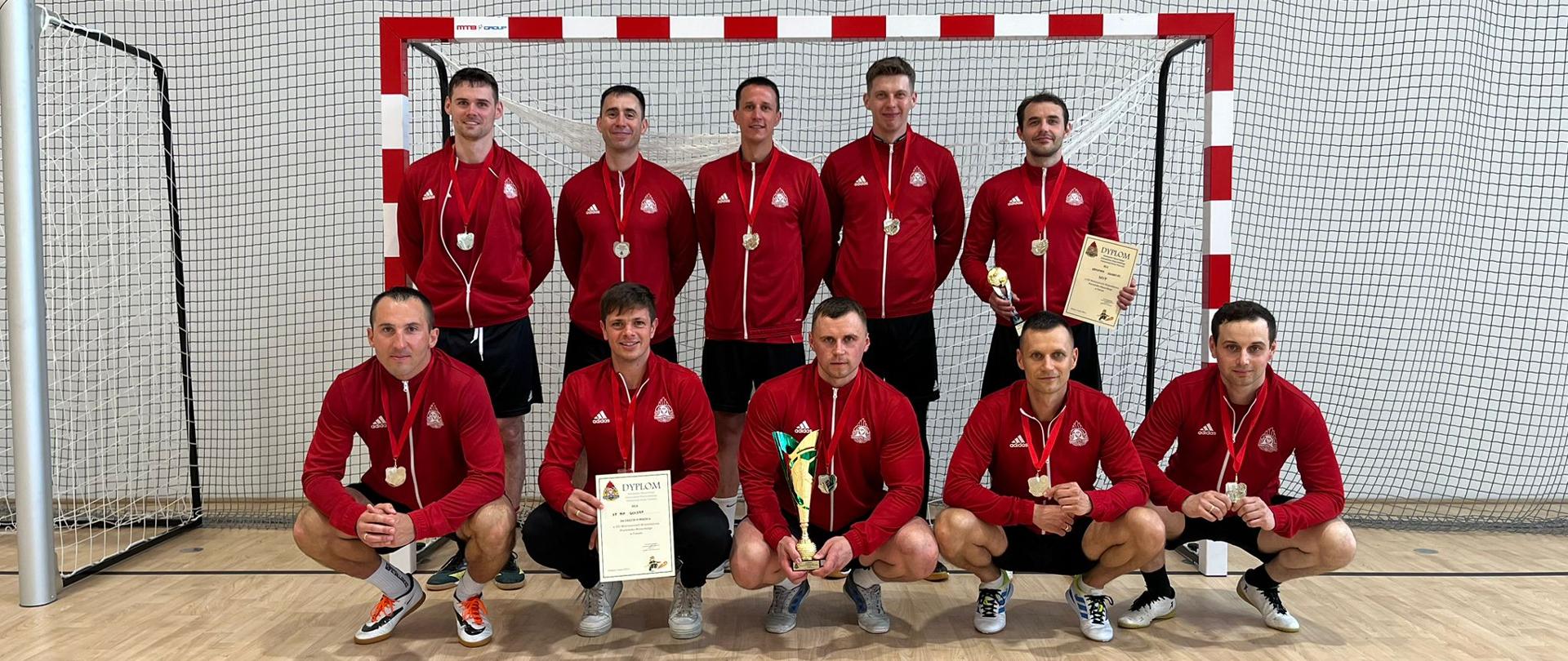 Zdjęcie przedstawia piłkarzy Komendy Powiatowej Państwowej Straży Pożarnej w Gołdapi z pucharami, medalami i dyplomami zlokalizowanych na tle bramki w hali sportowej po zakończonych XXI Mistrzostwach Województwa Warmińsko-Mazurskiego Strażaków Państwowej Straży Pożarnej w Futsalu