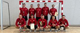 Zdjęcie przedstawia piłkarzy Komendy Powiatowej Państwowej Straży Pożarnej w Gołdapi z pucharami, medalami i dyplomami zlokalizowanych na tle bramki w hali sportowej po zakończonych XXI Mistrzostwach Województwa Warmińsko-Mazurskiego Strażaków Państwowej Straży Pożarnej w Futsalu