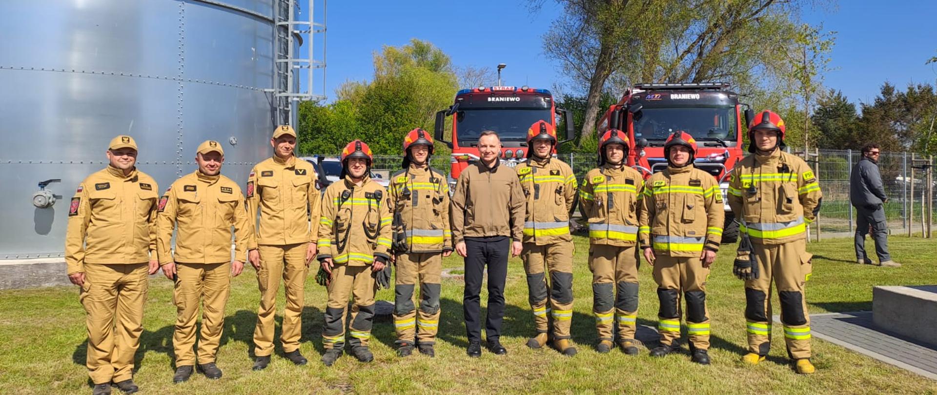 Na zdjęciu po środku stoi Prezydent Andrzej Duda. Obok niego z prawej i lewej strony strażacy w jasnych mundurach. Słonecznie. W tle samochody strażackie.