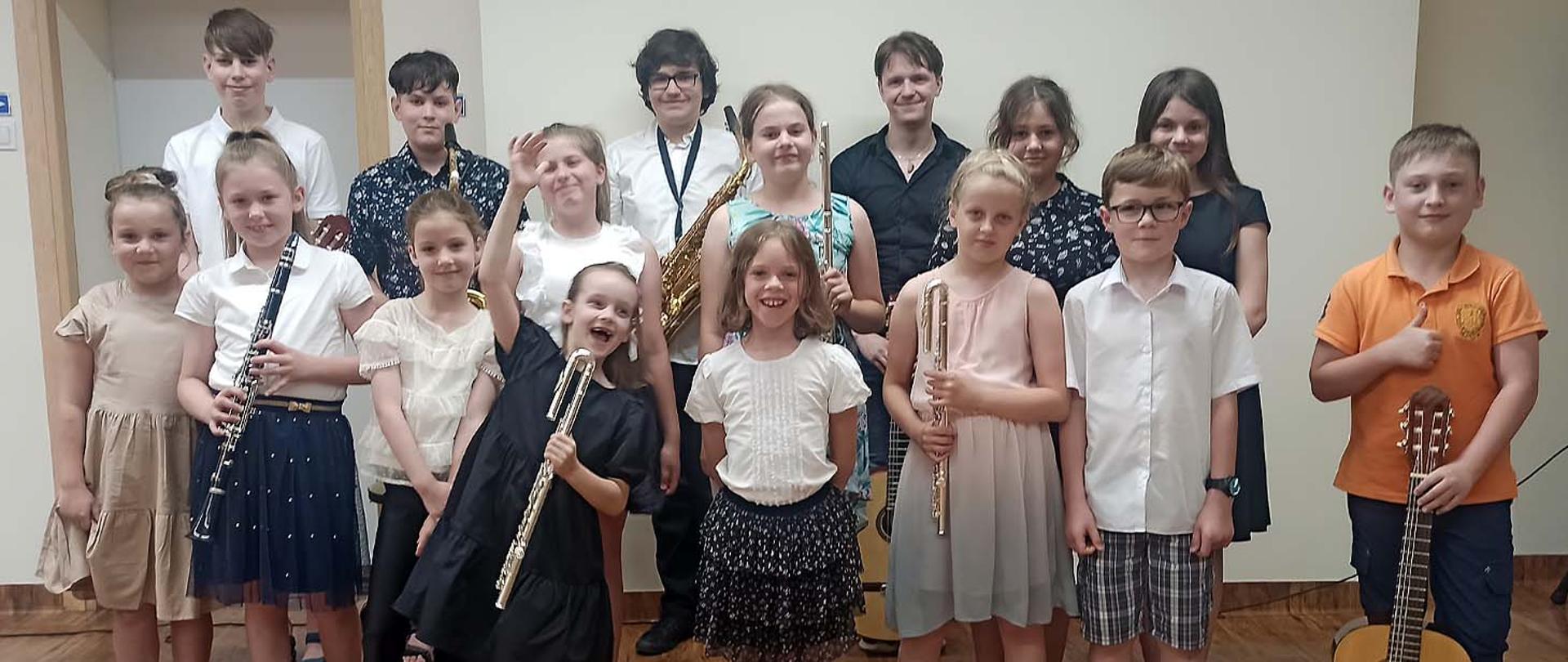 Uczniowie występujący podczas koncertu pozują do zdjęcia, stoją w auli szkoły z instrumentami w rękach.