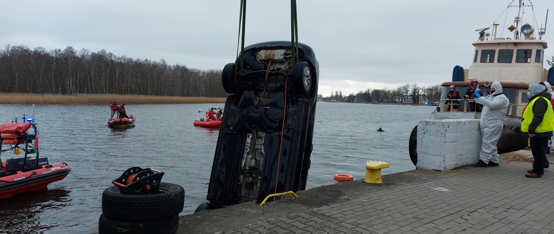 Na zdjęciu widać samochód wyciągany z wody przez dźwig. Na wodzie znajdują się trzy łodzie pontonowe straży pożarnej. po prawej stronie widać zacumowaną łódź.