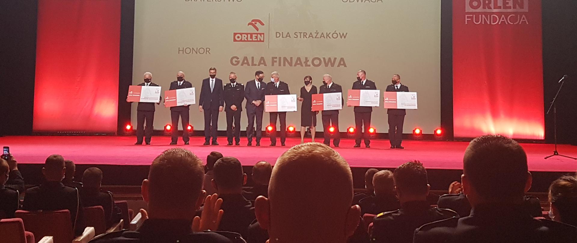 Finałowa Gala "Orlen dla Strażaków" na scenie stojący Premier, Komendant Główny, Prezes PKN Orlen wraz z innymi Komendantami, na widowni siedzą pozostali funkcjonariusze oraz druhowie OSP.