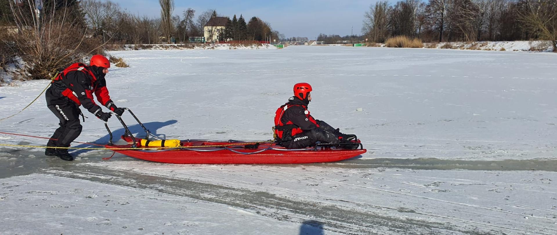 JRG Legionowo ćwiczenia z ratownictwa wodno - lodowego. Na zdjęciu zamarznięty akwen wodny z wyciętym przeręblem. 2 ratowników jeden pcha sanie lodowe drugi siedzi na saniach i podejmuje pozoranta. Wszyscy ubrani w skafandry lodowe.
