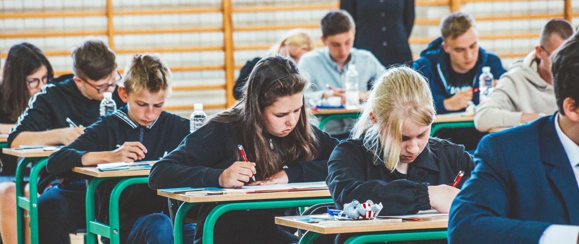 Dzieci i młodzież pochylona nad ławkami, rozwiązująca testy pisemne.