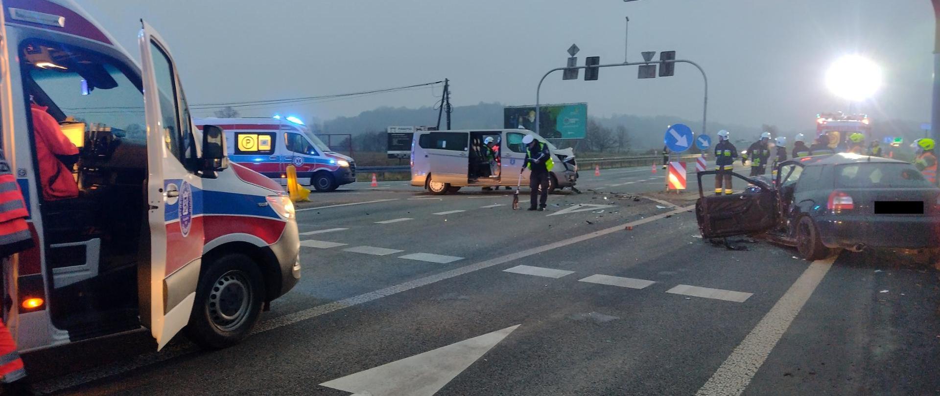 zdjęcie przedstawia skrzyżowanie drogowe, dwa pojazdy biorące udział w zdarzeniu, dwie karetki pogotowia, funkcjonariuszy Straży Pożarnej oraz Policjanta