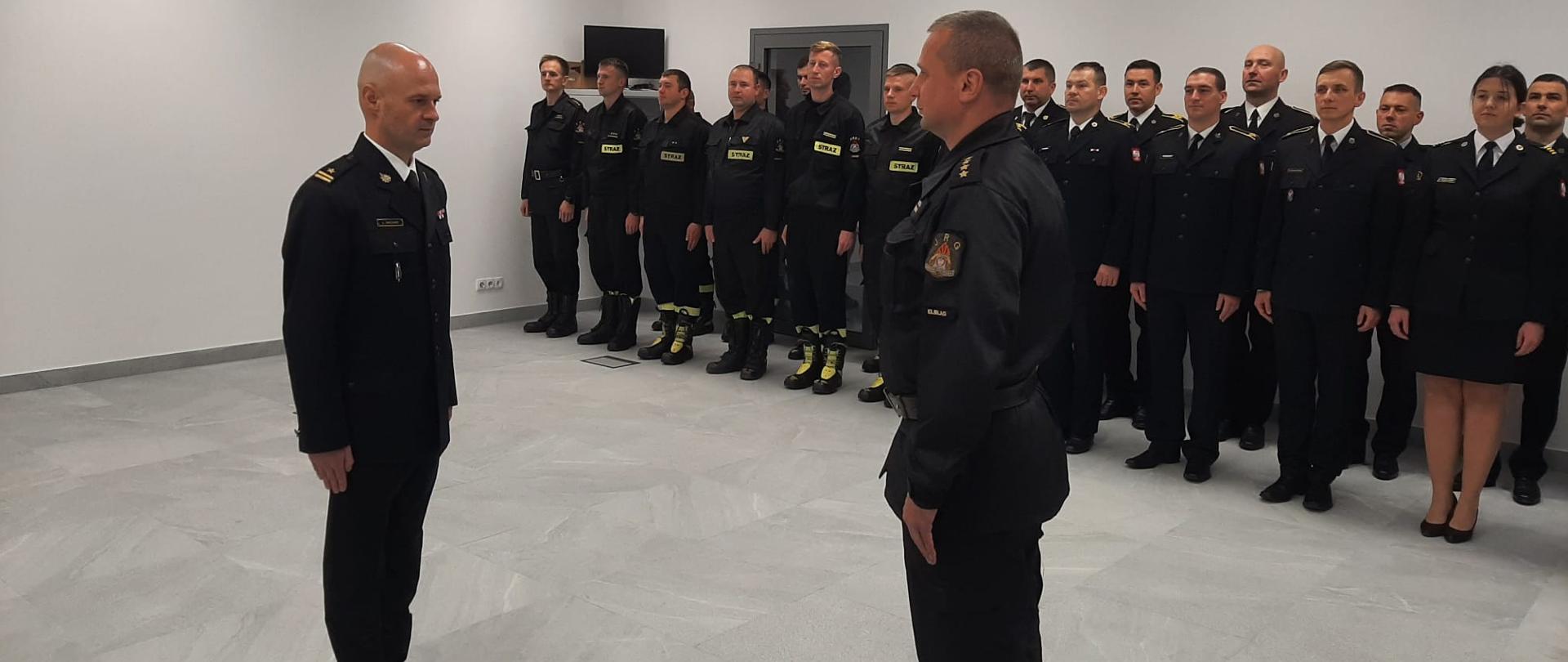 Na pierwszym planie dowódca jednostki składa meldunek komendantowi miejskiemu. W tle w dwuszeregu stoją w mundurach strażacy.