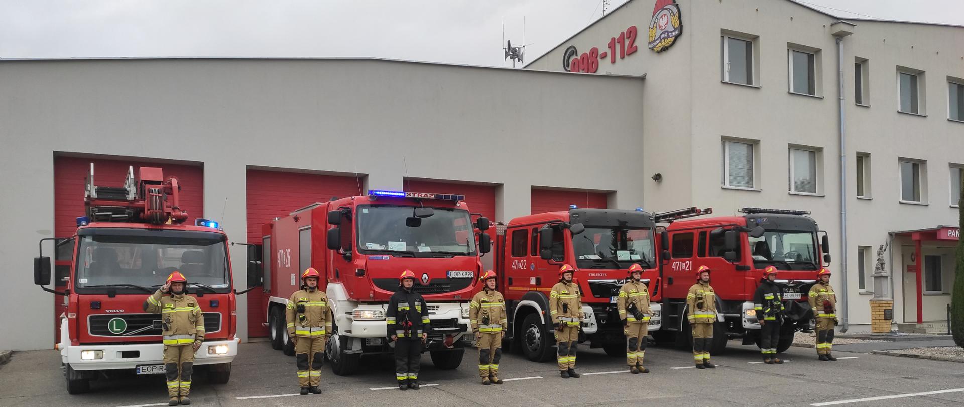 Zdjęcie przedstawia funkcjonariuszy KP PSP w Opocznie w ubraniach specjalnych oddających honory. W tle znajdują się Pojazdy bojowe z włączona sygnalizacją świetlną oraz budynek Komendy.