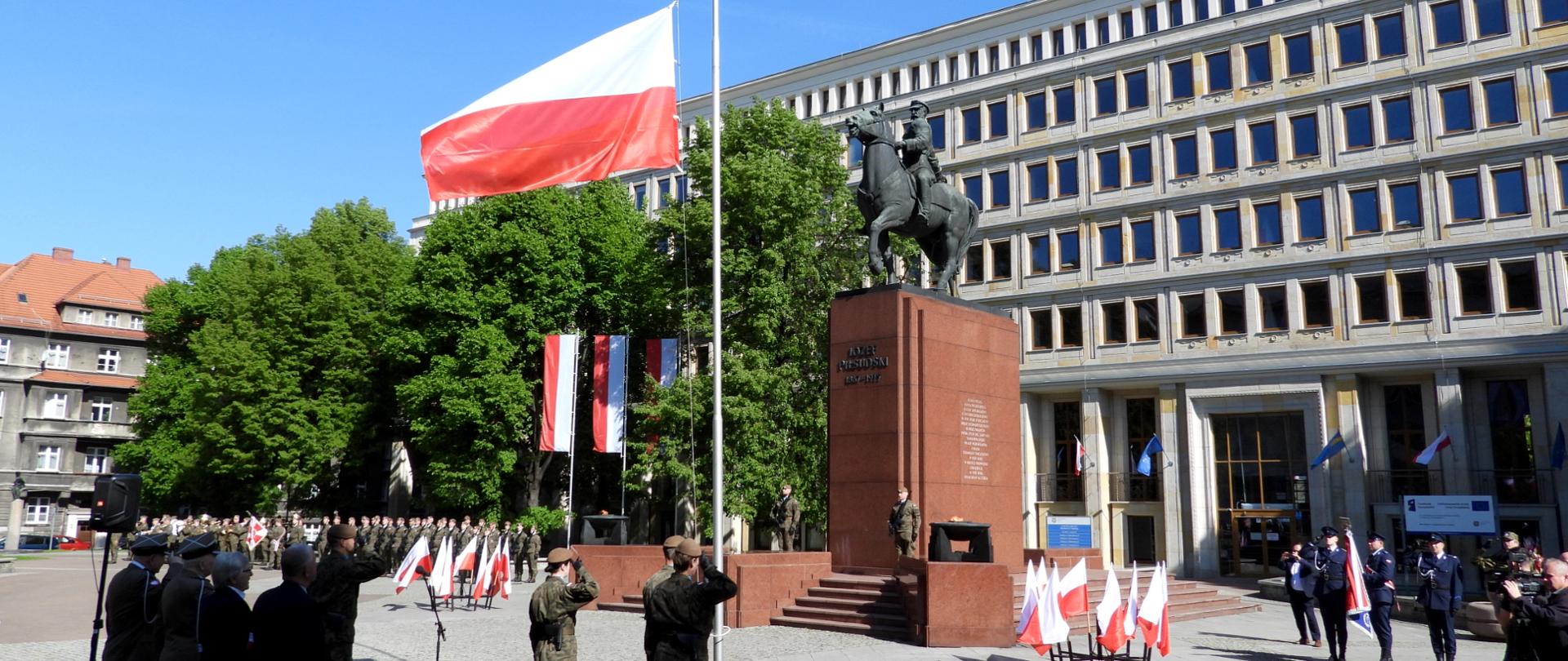 Zdjęcie przedstawia oficjalną uroczystość państwową odbywającą się na placu miejskim pod gołym niebem, w słoneczny dzień. W centrum kadru, na wysokim maszcie, unosi się flaga Polski – biało-czerwona, rozwinięta przez wiatr. Pod masztem kilku żołnierzy w mundurach salutuje. Na prawo od masztu znajduje się monumentalny pomnik przedstawiający postać Józefa Piłsudskiego siedzącego na koniu. Pomnik stoi na wysokim, czerwonobrązowym cokole z wyrytym napisem: "KAZIMIERZ PUŁASKI MARSZAŁEK".W tle, za pomnikiem, znajduje się duży, nowoczesny budynek z licznymi kwadratowymi oknami – prawdopodobnie gmach urzędu lub instytucji publicznej. Na placu zgromadzony jest tłum uczestników: żołnierze w szyku, funkcjonariusze w wyjściowych mundurach osoby w garniturach, a także media z kamerami. Widać również ustawione biało-czerwone flagi oraz mikrofon na statywie.