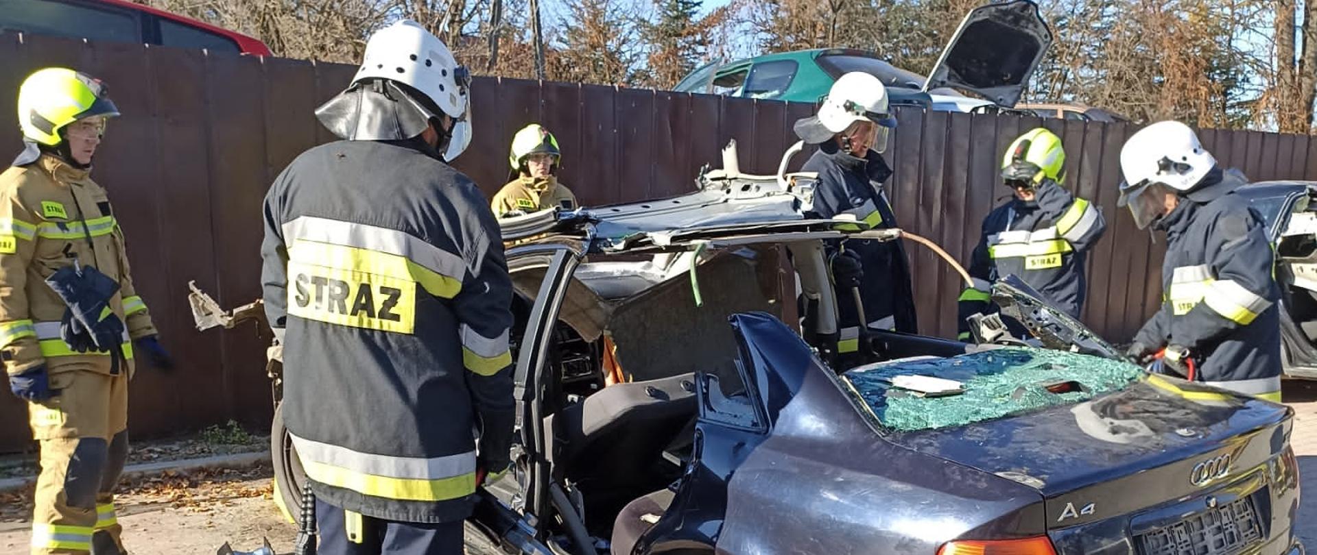 Kolorowa fotografia wykonana w słoneczny dzień przedstawia kilku umundurowanych na bojowo strażaków, którzy ćwiczą przy wraku samochodu osobowego.