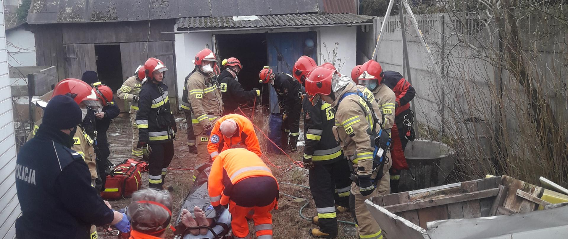 Zdjęcie przedstawia teren akcji - na noszach widać wyciągniętego ze studni mężczyznę. Po prawej stronie znajduje się studnia. Nad mężczyzną pochylają się ratownicy z Pogotowia Ratunkowego. Wokół noszy stoją strażacy oraz Policja.