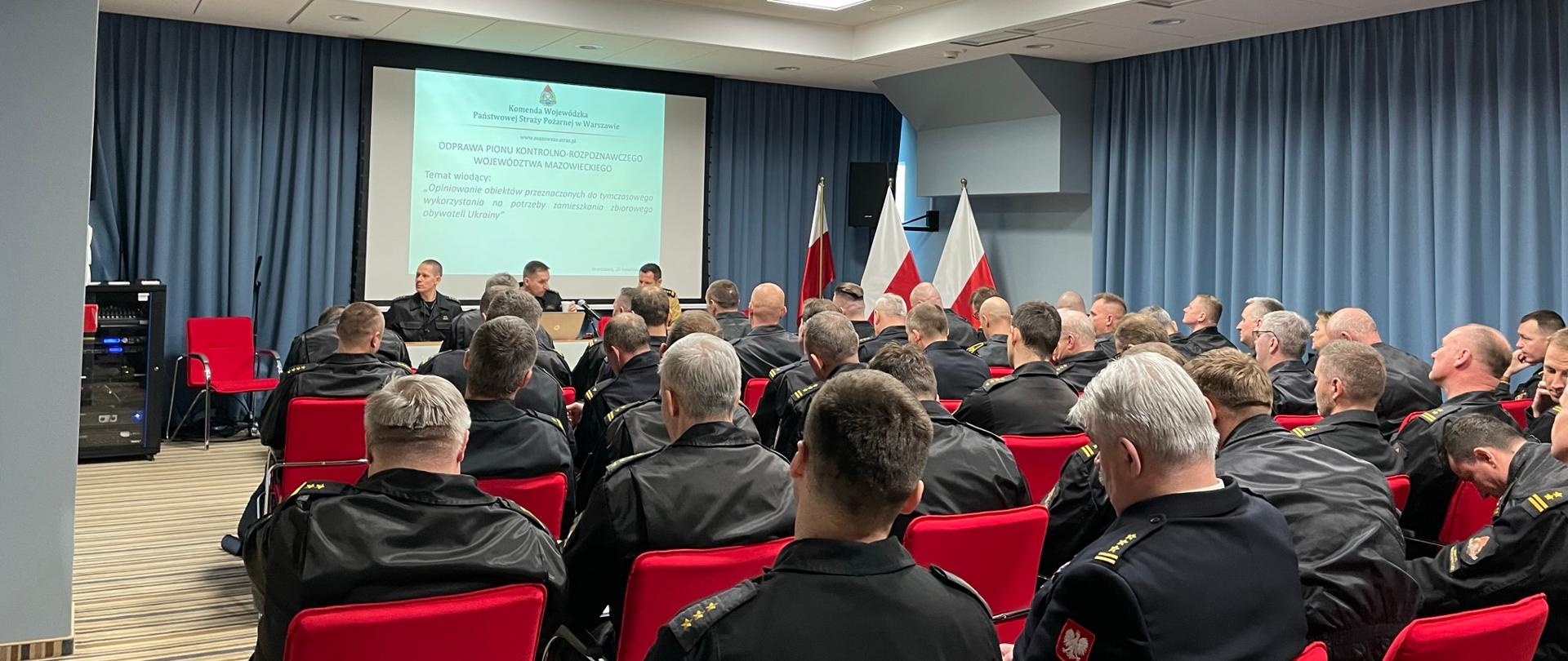 odprawa pionu kontrolno-rozpoznawczego PSP województwa mazowieckiego. uczestnicy siedzą na krzesłach w sali konferencyjnej. na przodzie przy mównicy siedzi zastępca mazowieckiego komendanta PSP oraz naczelnicy wydziału kontrolno-rozpoznawczego komendy wojewódzkiej psp w warszawie. w tle wyświetlana jest prezentacja na rzutniku.