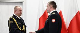 Strażak w mundurze wyjściowym ze sznurem podaje dłoń strażakowi w mundurze wyjściowym trzymającym teczkę za mężczyznami ustawione są trzy flagi Polski oraz znajdują się drzwi.
