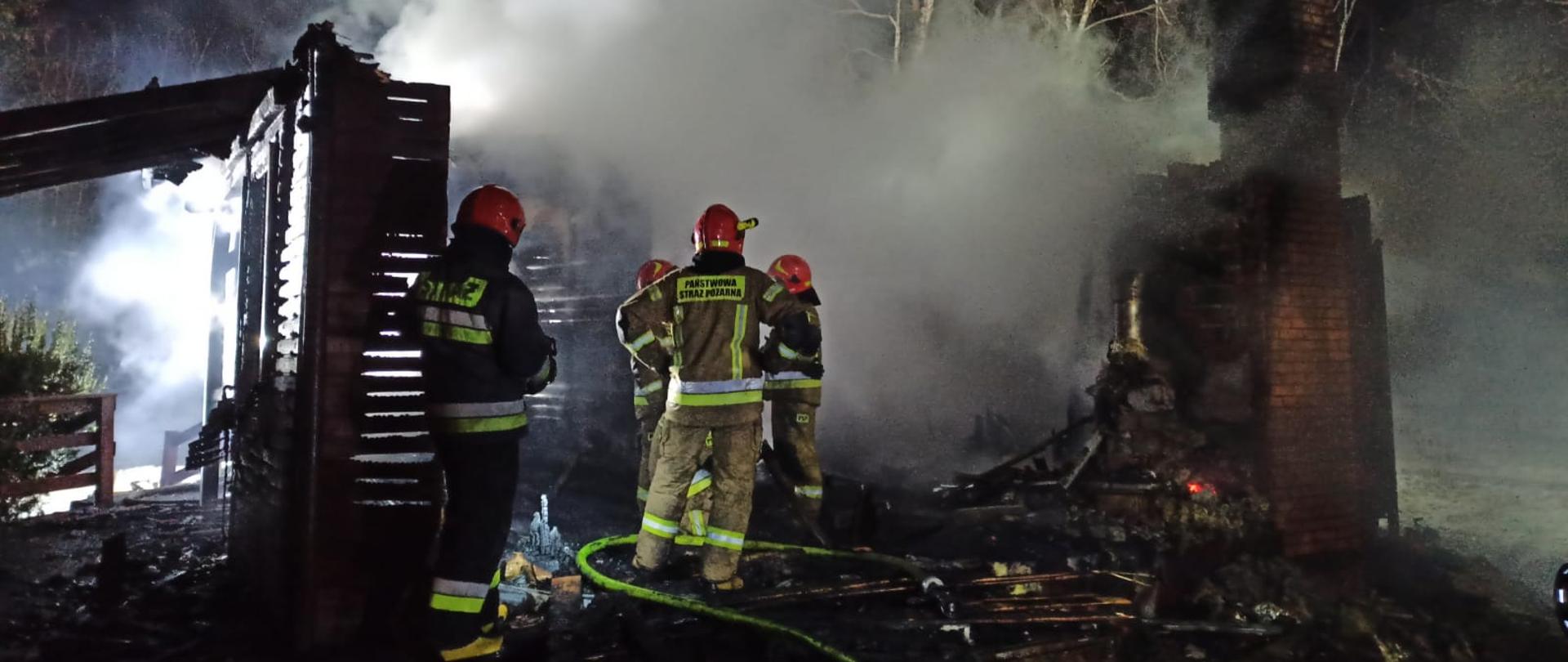 Zdjęcie przedstawia częściowo spalony domek letniskowy. Na jego terenie stoją strażacy poubierani w ubrania specjalne koloru żółtego i granatowego. Na głowach mają czerwone hełmy. Na ziemi leżą rozwinięte węże gaśnicze w kolorze żółtym. Ze spalonych elementów budynku unosi się jeszcze dym i para wodna. Zdjęcie wykonane w porze nocnej. 