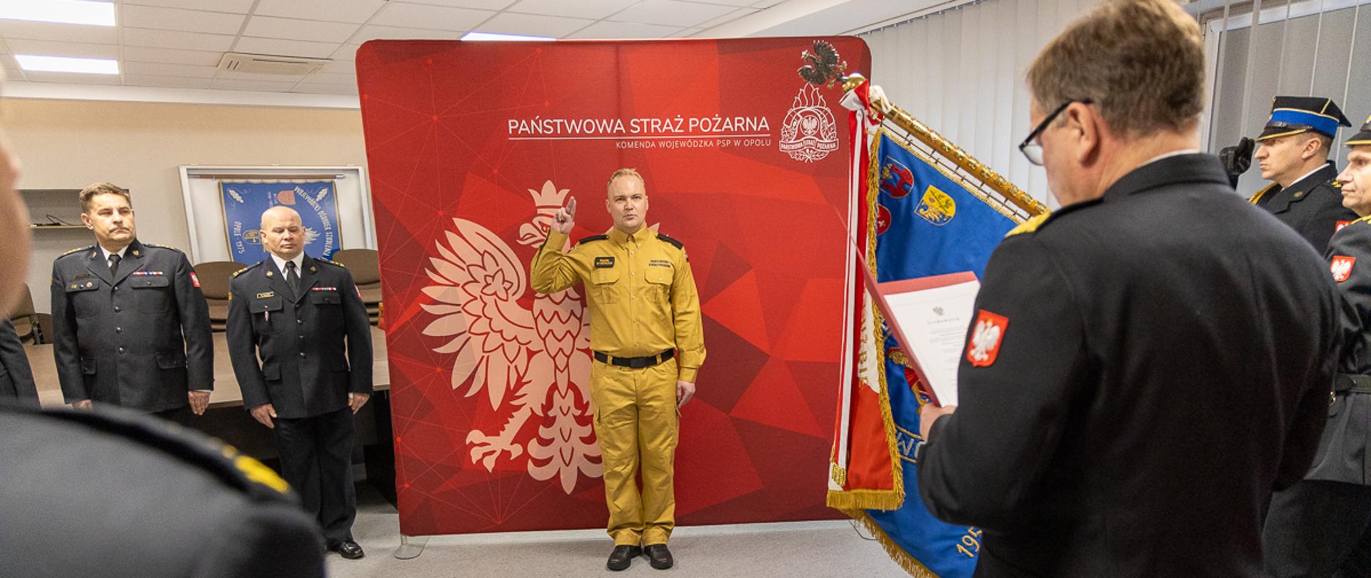 Uroczystość ślubowania strażaka Państwowej Straży Pożarnej. Opolski Komendant Wojewódzki PSP odczytuje treść ślubowania. W tle funkcjonariusze Komendy Wojewódzkiej PSP w Opolu, poczet sztandarowy Komendy Wojewódzkiej PSP w Opolu. Na pierwszy planie ślubujący strażak w umundurowaniu służbowym na tle czerwonego banneru Państwowej Straży Pożarnej.