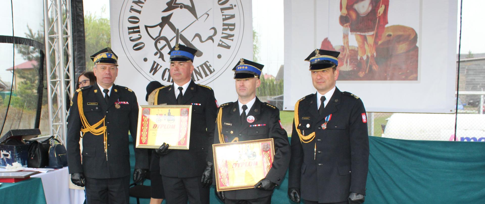 czterech strażaków stoi pod namiotem, dwóch z nich trzyma ramki z dyplomem komendanta głównego PSP